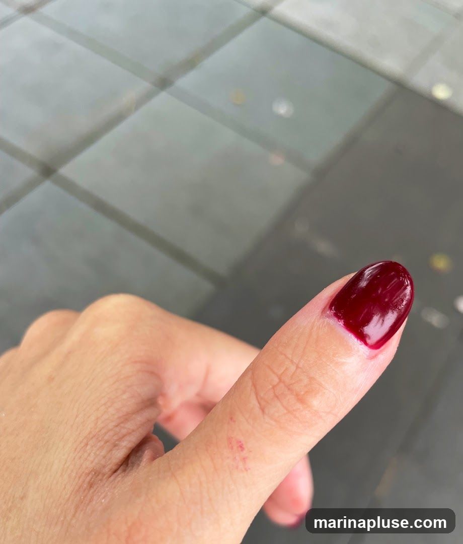A close-up shot of a thumb with dark red nail polish. The nail polish is glossy and covers most of the nail, with a small unpolished area near the cuticle. There are a few small red marks on the skin of the thumb. The background appears to be a tiled surface, possibly outdoors.