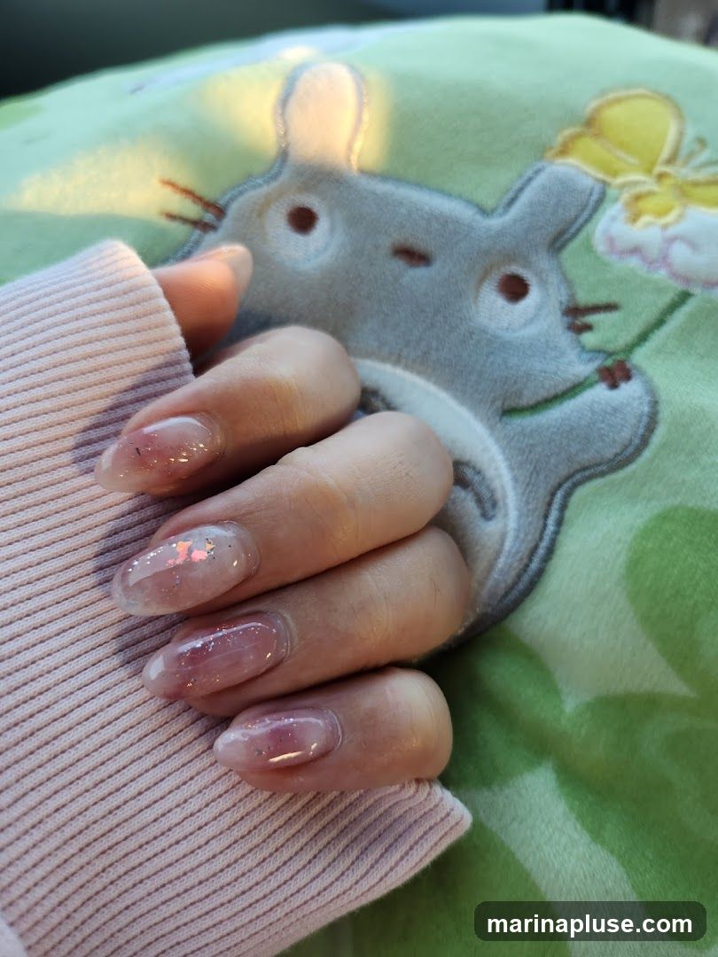 A close-up shot of a person's hand with light pink, glittery, oval-shaped nails. The hand is resting on a light pink ribbed fabric, possibly a sweater. In the background, a light green pillow with a large embroidered image of the character Totoro is visible.