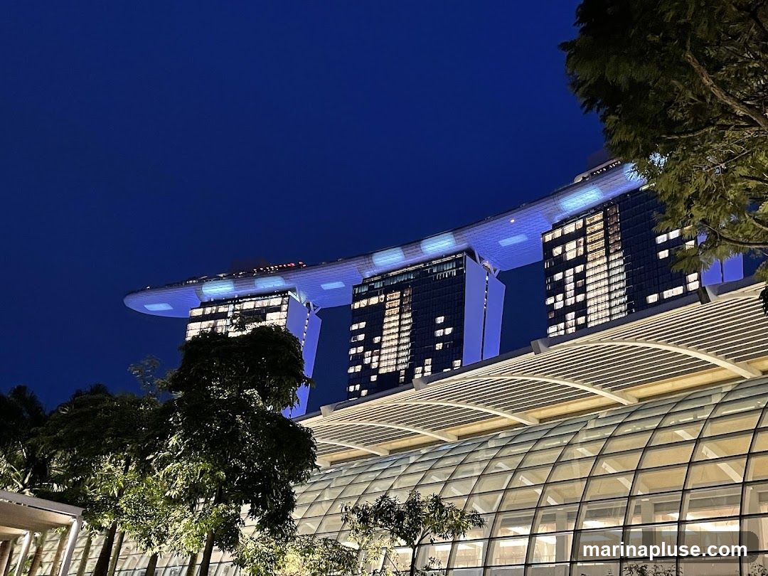 A nighttime view of the Marina Bay Sands hotel in Singapore, with its iconic three towers connected by a sky park. The building is illuminated with blue lights, and the windows of the towers show warm interior lighting. In the foreground, a modern glass-covered structure with a curved roof is visible, along with trees and foliage.