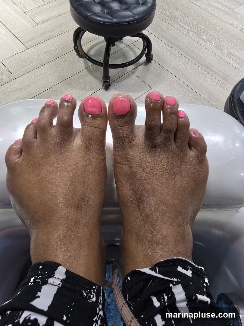 A close-up view of a person's feet with bright pink toenail polish, resting in a pedicure chair. A black, tufted stool is visible in the background, along with light-colored wood-grain flooring.
