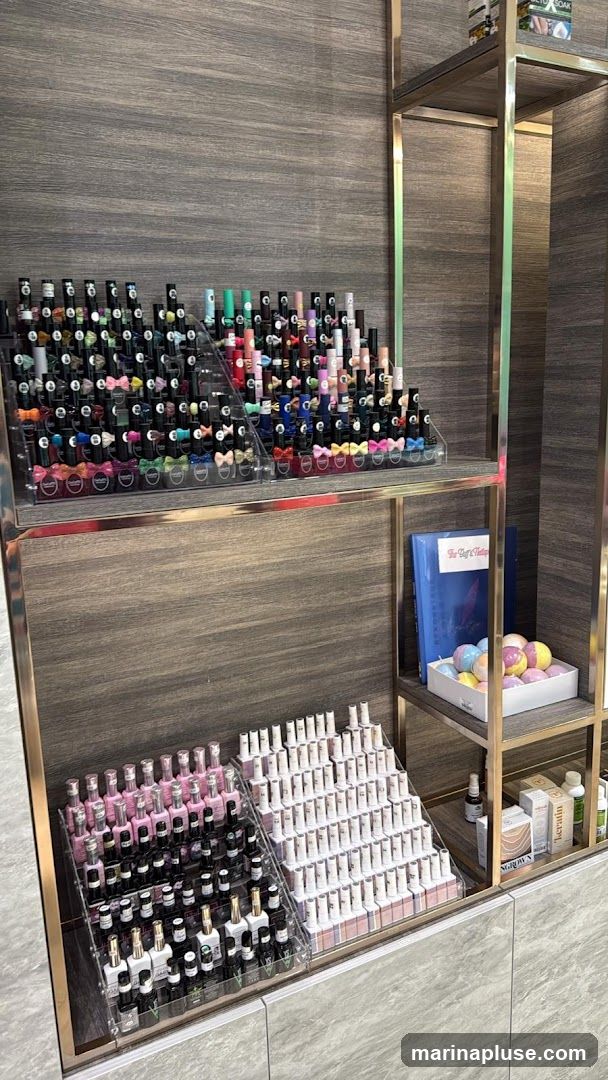 A display shelf filled with numerous bottles of nail polish in various colors. The polishes are arranged on tiered acrylic stands. On a higher shelf, there are bath bombs and a book. On the right side, there are boxes of skincare or nail care products.