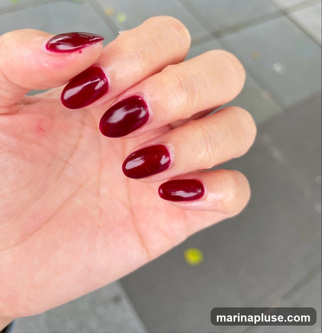 A close-up shot of a person's left hand, showcasing their nails which are painted in a deep, glossy dark red color. The nails are shaped into a rounded oval. The background is blurred and appears to be an outdoor setting with a grey, tiled surface.