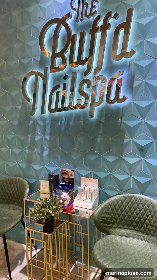 A close-up view of the interior of The Buff'd Nailspa, featuring a prominent metallic sign with the salon's name against a textured teal wall. In the foreground, a small gold-accented table displays various nail care products and a decorative plant. Two plush green chairs are visible on either side of the table.