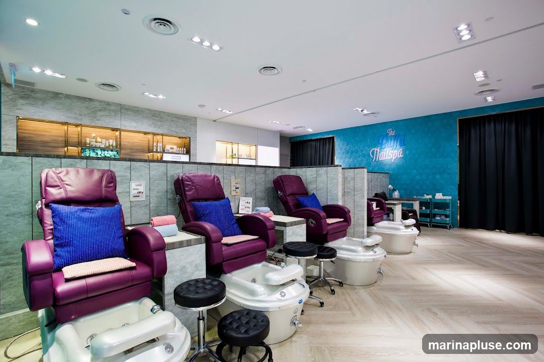 A well-lit nail spa with multiple pedicure stations. Each station features a plush purple reclining chair with a blue decorative pillow and a white foot bath. Stools are placed in front of the chairs. In the background, shelves display various beauty products. The spa's name, "The Royal Nailspa," is visible on a teal, hexagonal-patterned wall. Black curtains hang on the right side of the room.