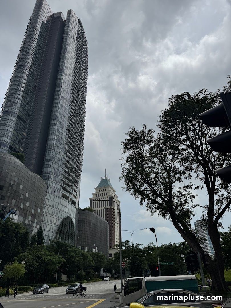 A view of a city street with a tall, modern skyscraper on the left and a more traditional, multi-story building in the background. Trees line the street, and traffic signals and vehicles are visible. The sky is overcast with clouds.