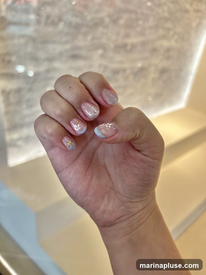 A close-up shot of a person's hand displaying a set of short, natural-looking nails adorned with delicate nail art. The design features a sheer pink base with subtle white floral patterns, glitter accents, and small gold embellishments. The background is softly blurred, suggesting an indoor setting with warm lighting.