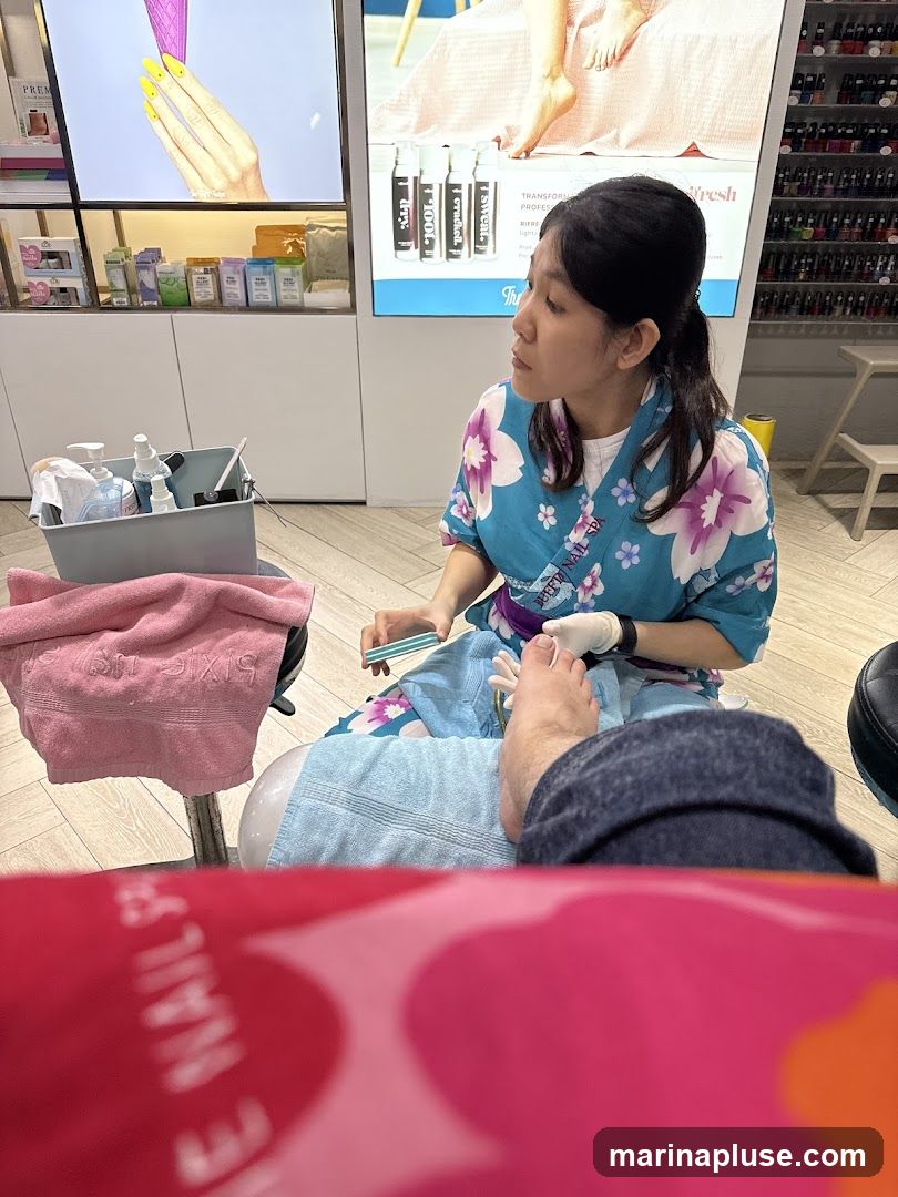 A nail technician in a blue floral robe is filing a client's toenail. The client's foot is resting on a blue towel. In the background, there are shelves stocked with nail polish bottles and displays showing nail art and beauty products. A pink towel is draped over a chair in the foreground.