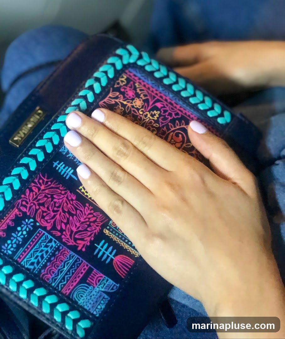 A close-up shot shows a person's hand with light purple nail polish resting on a dark blue bag. The bag features a colorful, embroidered pattern with floral and geometric designs in shades of pink, orange, blue, and teal. The hand is positioned over the central part of the bag, obscuring some of the design. The person is wearing blue denim clothing.