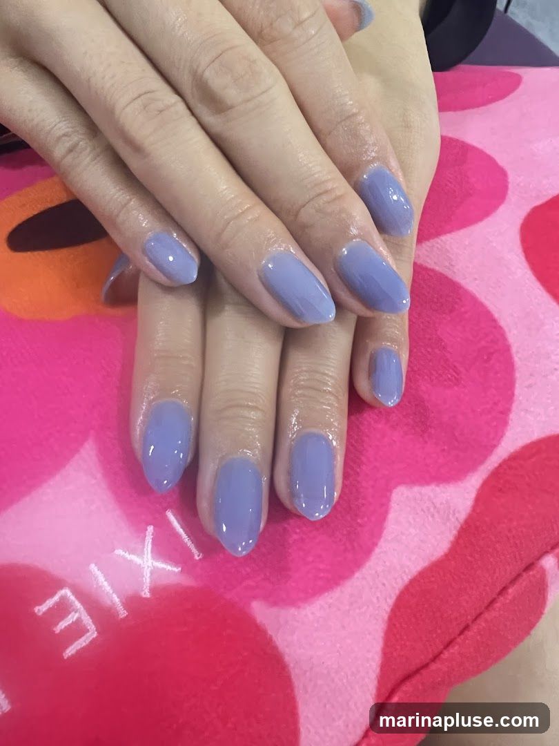 A close-up shot of a person's hands with freshly painted lavender-colored almond-shaped nails. The nails are glossy and well-manicured. The hands are resting on a pink and red patterned surface, possibly a towel or blanket, with the word "IXIE" visible in white lettering.