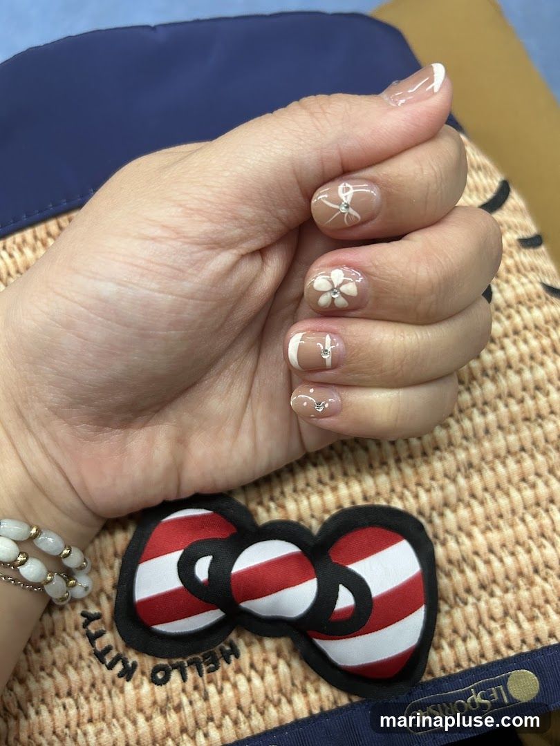 A close-up shot of a hand with manicured nails featuring a light brown base with white floral designs and small rhinestones. The nails also have a French tip accent. In the foreground, a Hello Kitty bow with red and white stripes is visible, along with the text "HELLO KITTY" and "LESPORTSAC" on a textured background.