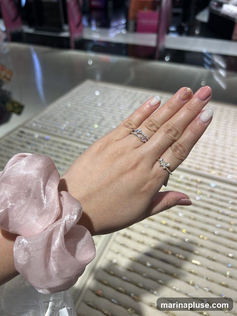 A hand with two rings on display, with a pink scrunchie on the wrist. The background shows rows of jewelry on display stands, suggesting a retail environment. The nails are painted in various pastel shades and glitter.