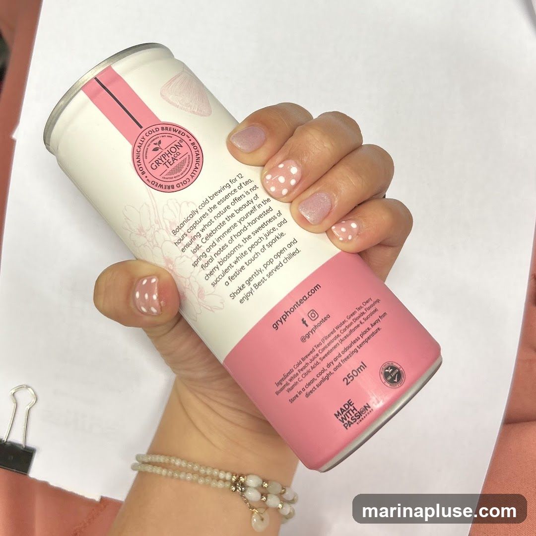 A hand with white polka dot nail art holds a can of Gryphon Tea. The can is white with pink accents and features floral illustrations and text describing the tea's flavor profile and ingredients. The text indicates it is a cold-brewed tea with cherry blossom and white peach flavors.