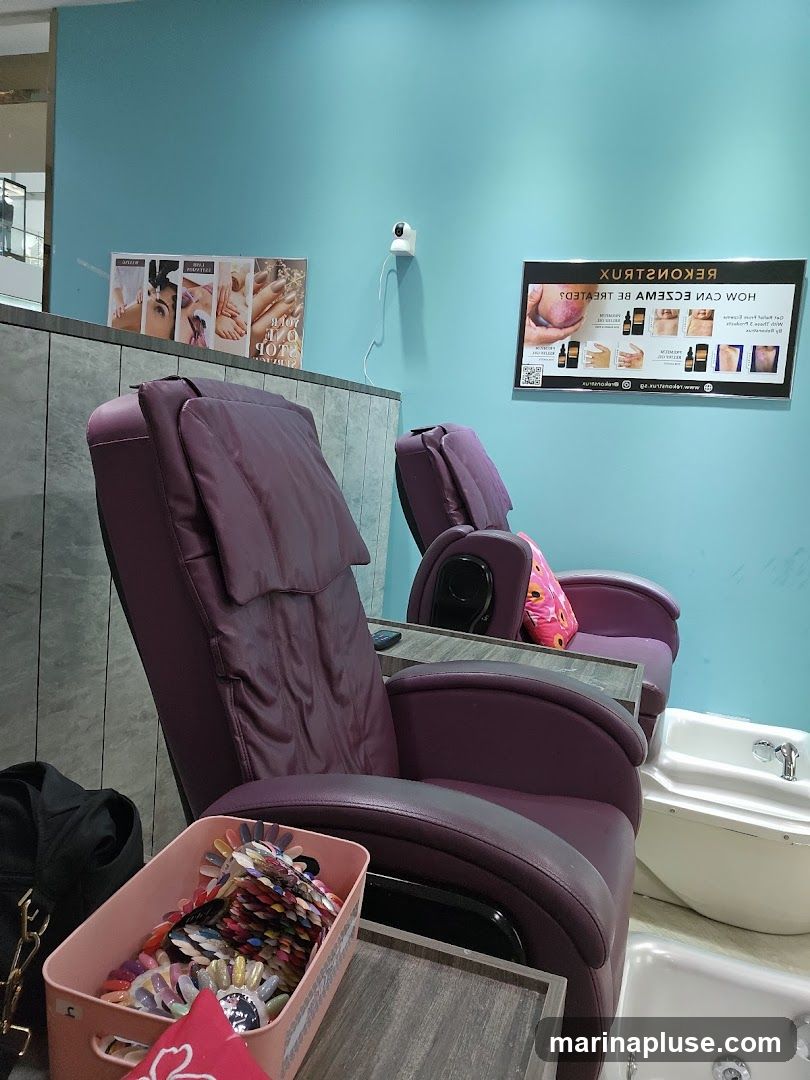 A nail salon interior with two purple pedicure chairs facing white foot baths. A pink bin filled with nail polish color samples sits in the foreground. On the wall, there are two framed posters: one with images of manicures and pedicures, and another advertising a product for eczema treatment. A security camera is mounted on the teal-colored wall.