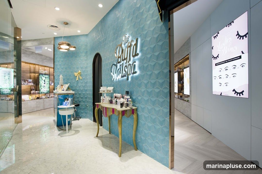 The interior of a modern nail spa with teal textured walls and a neon sign that reads "The Buffd Nailspa". There are display shelves with products, a decorative table with more products, and a poster on the wall advertising lash and brow services. The floor is tiled with a marble pattern.