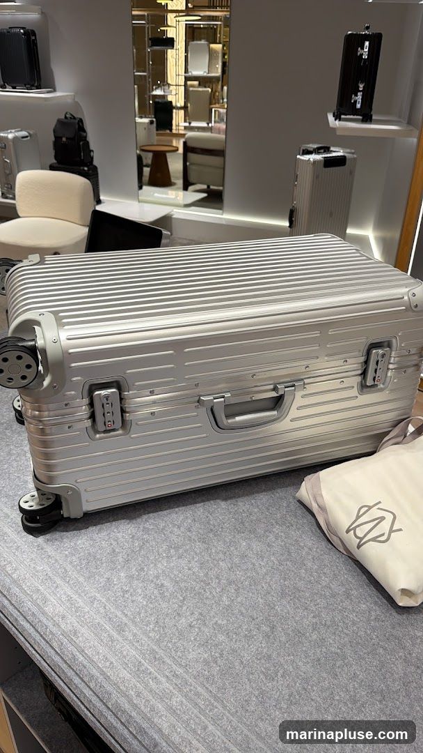 A large, silver, ribbed suitcase with black wheels and combination locks is displayed prominently on a grey carpeted surface. In the background, other luggage pieces, including black and silver suitcases and a black backpack, are visible on shelves and a chair. A mirror reflects more of the store's interior, showing additional luggage and display shelves. A light-colored dust bag with a logo is placed next to the large suitcase.