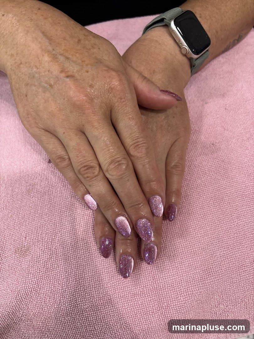 A close-up shot of a person's hands with freshly manicured nails. The nails are almond-shaped and painted with a shimmering pink glitter polish. The hands are resting on a pink textured towel. A smartwatch with a grey band is visible on the left wrist.