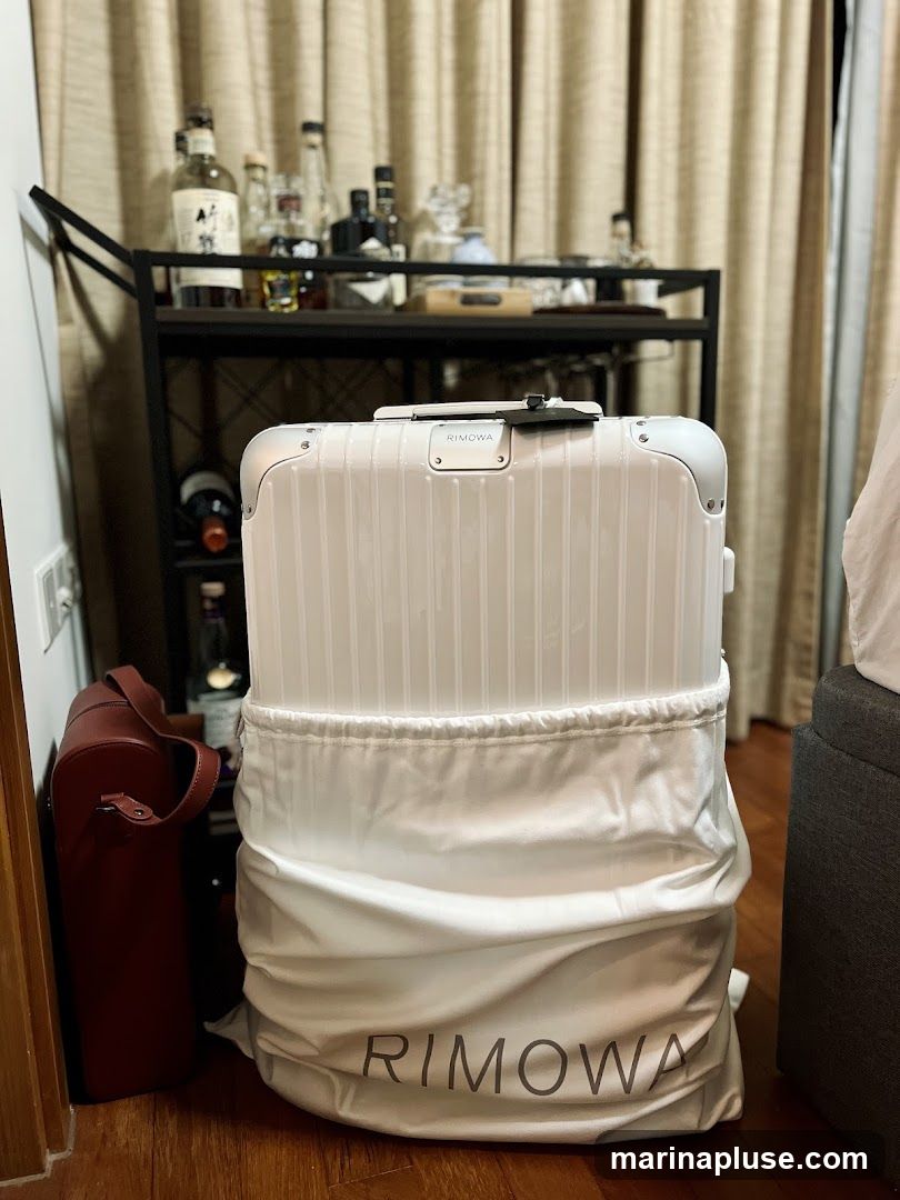 A white Rimowa suitcase is covered by a white protective bag with the Rimowa logo. To the left of the suitcase is a dark red cylindrical bag. In the background, a bar cart holds various bottles of alcohol and glassware. Curtains are visible behind the bar cart.