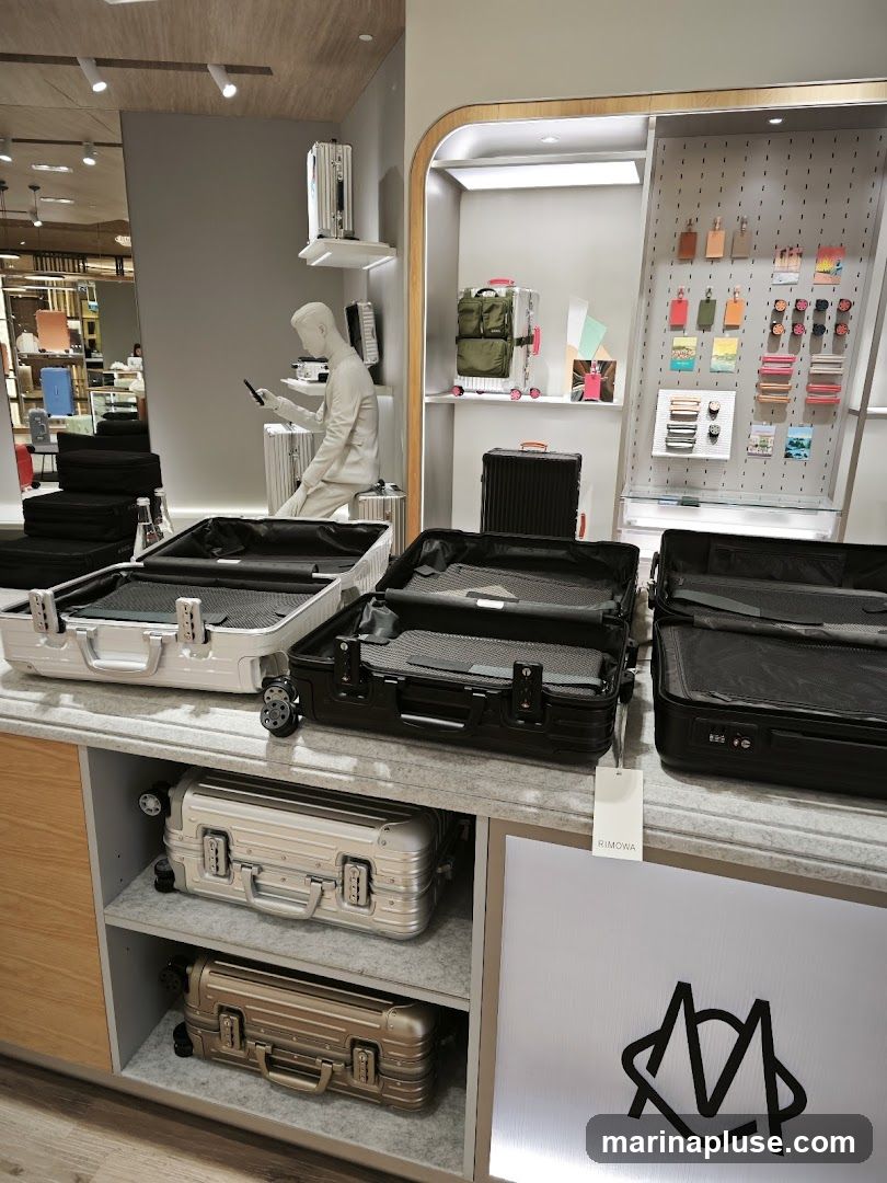 A display of various luggage pieces, including open suitcases and smaller travel accessories. A mannequin is seated, holding a phone. The brand name 'RIMOWA' is visible on a tag.
