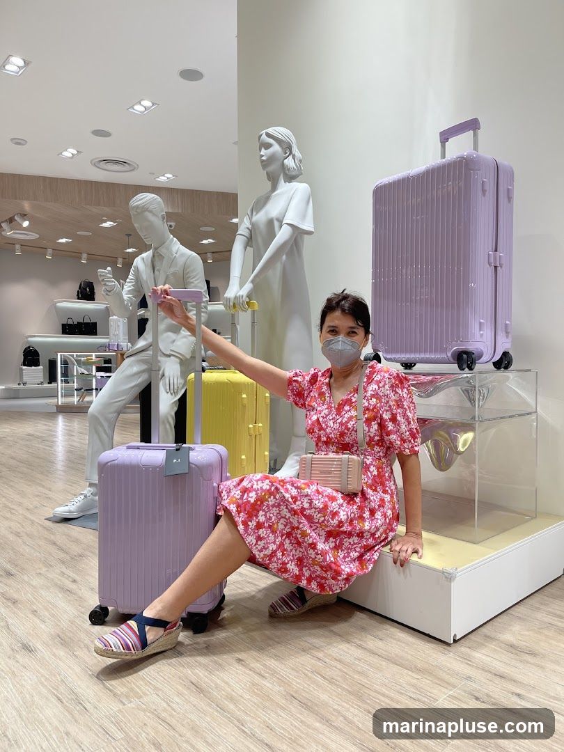 A woman wearing a floral dress and a face mask poses in a store with several mannequins and luggage displays. She is seated next to a light purple suitcase, with her hand on its handle. Behind her, a yellow suitcase and a larger light purple suitcase are displayed on a platform. Two white mannequins, one male and one female, are also part of the display. The store interior is visible in the background, with shelves displaying bags and other merchandise.