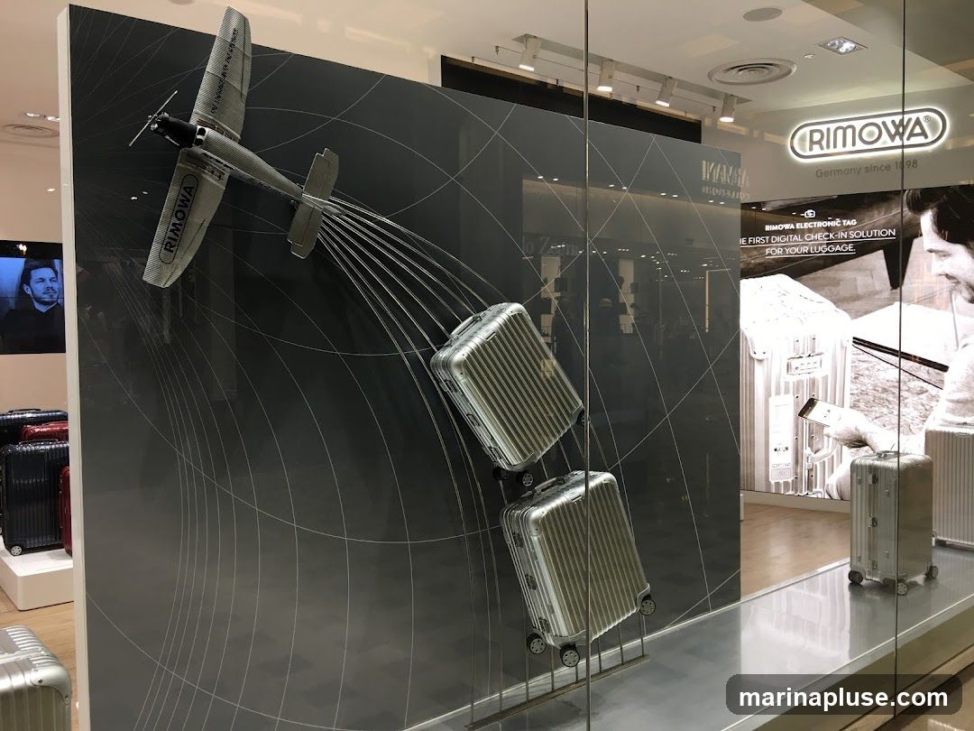 A display inside a Rimowa store features a model airplane suspended above several pieces of luggage. The airplane is connected to the luggage by thin wires, creating a visual effect of the plane pulling the bags. The store's branding, "RIMOWA Germany since 1898," is visible on the wall and in the window. A large advertisement for "RIMOWA ELECTRONIC TAG" is also displayed, highlighting it as "THE FIRST DIGITAL CHECK-IN SOLUTION FOR YOUR LUGGAGE."