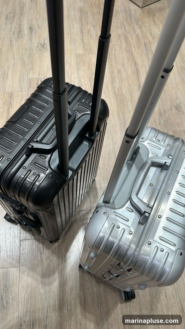 A close-up shot shows two suitcases, one black and one silver, with their telescopic handles extended. The suitcases are placed on a light-colored wooden floor. The black suitcase is on the left, and the silver one is on the right. Both suitcases have a ribbed exterior design and visible latches and wheels.