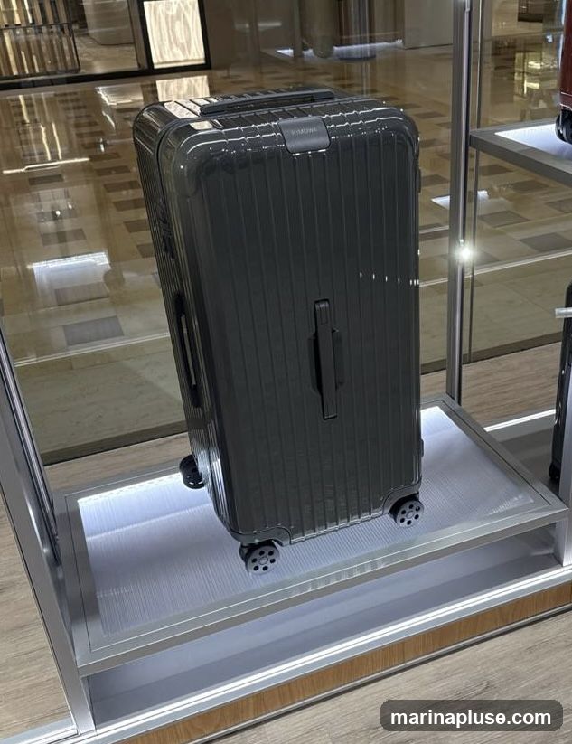 A dark gray, ribbed suitcase with the brand name RIMOWA visible on its top, is displayed on a shelf with integrated LED lighting. The suitcase has black wheels and handles. It is positioned within a display case, with reflections of the surrounding environment visible on the glass.