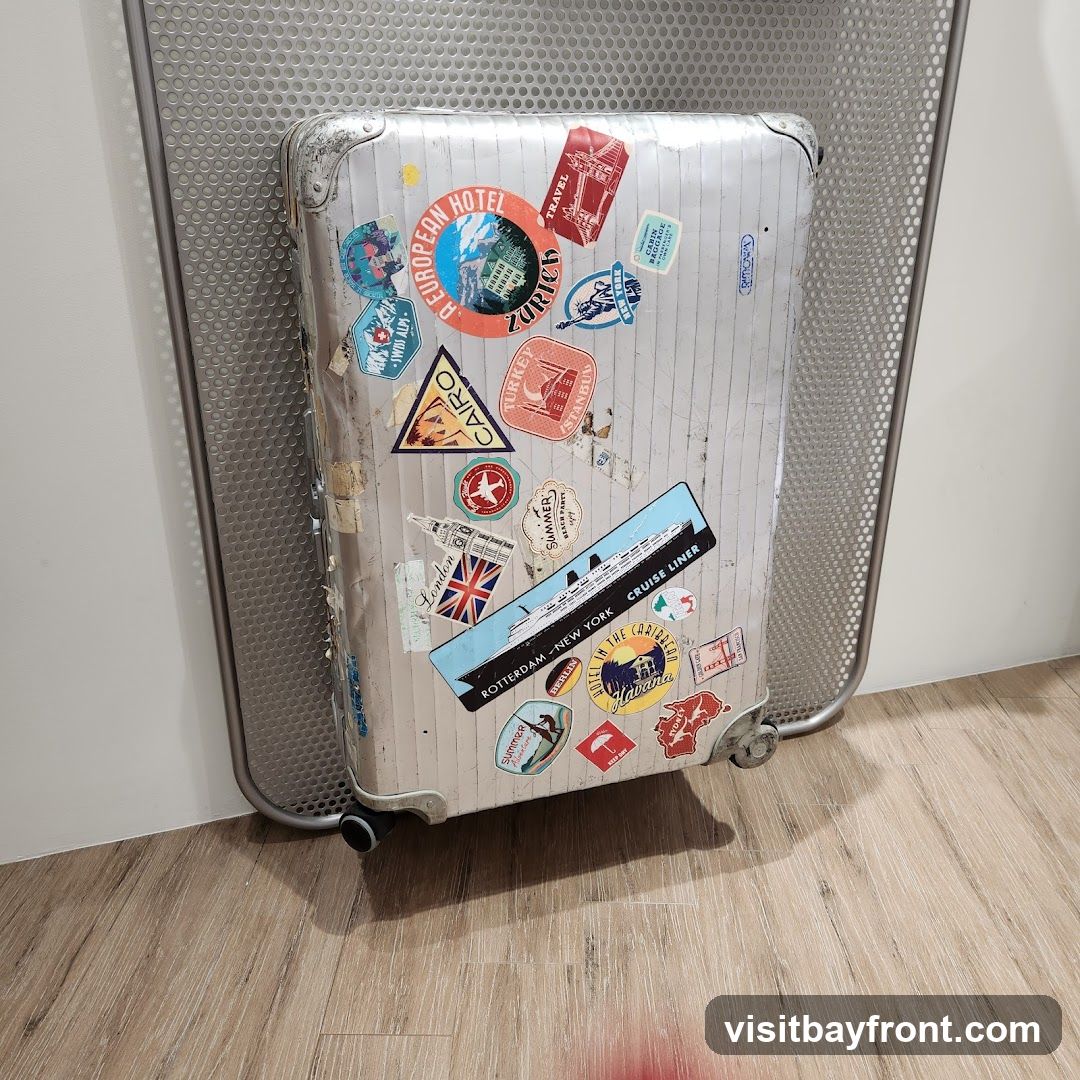 A vintage silver suitcase covered in various travel stickers from different destinations and modes of transport. Destinations include Zurich, Cairo, Turkey, London, Rotterdam, New York, and Havana. There are also stickers for a cruise liner and a Swiss Alps adventure. The suitcase is placed on a wooden floor next to a perforated metal screen.