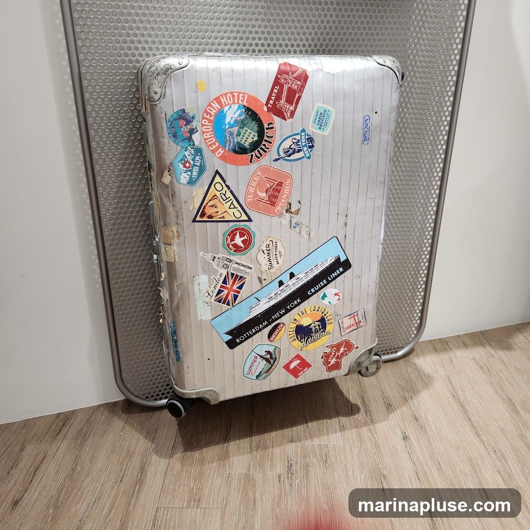 A vintage silver suitcase covered in various travel stickers from different destinations and modes of transport. Destinations include Zurich, Cairo, Turkey, London, Rotterdam, New York, and Havana. There are also stickers for a cruise liner and a Swiss Alps adventure. The suitcase is placed on a wooden floor next to a perforated metal screen.