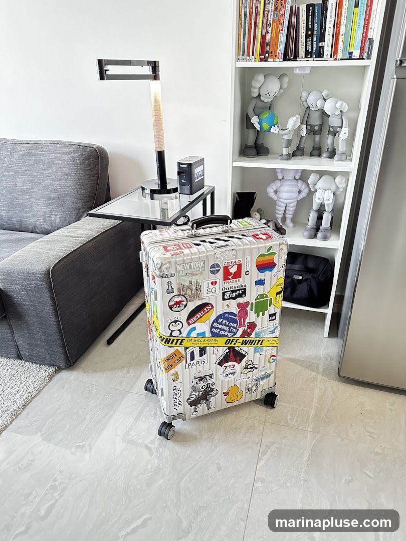 A silver suitcase covered in various stickers, including city names like New York, Paris, and Berlin, as well as cartoon characters and logos, stands on a tiled floor. To the left is a grey sofa and a modern black lamp on a side table. To the right, a white bookshelf is filled with books and displays several KAWS collectible figures. A small black printer is also visible on the side table.
