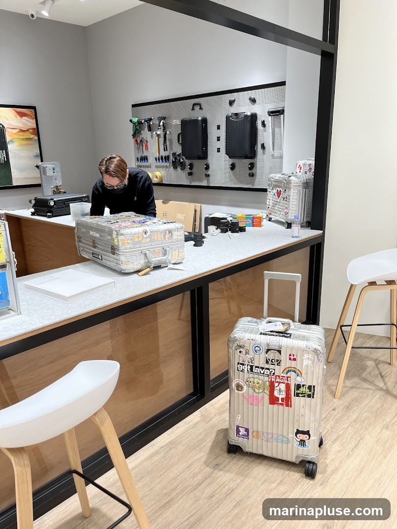 An interior view of a store with luggage on display. A person is behind a counter, and several suitcases are visible, including one with many stickers. Tools and two black suitcases are mounted on a pegboard on the wall. Two white stools are in the foreground.