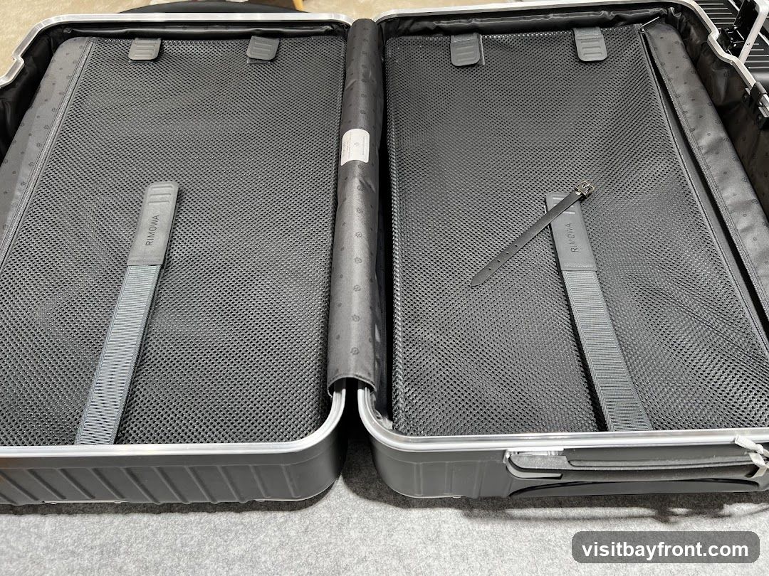 An open Rimowa suitcase, showing the interior compartments. The suitcase is dark gray with silver trim. The interior is lined with black mesh fabric and features two gray straps with the "RIMOWA" logo printed on them. One strap is on the left side, and the other is on the right side, secured with a buckle. The suitcase appears to be empty.