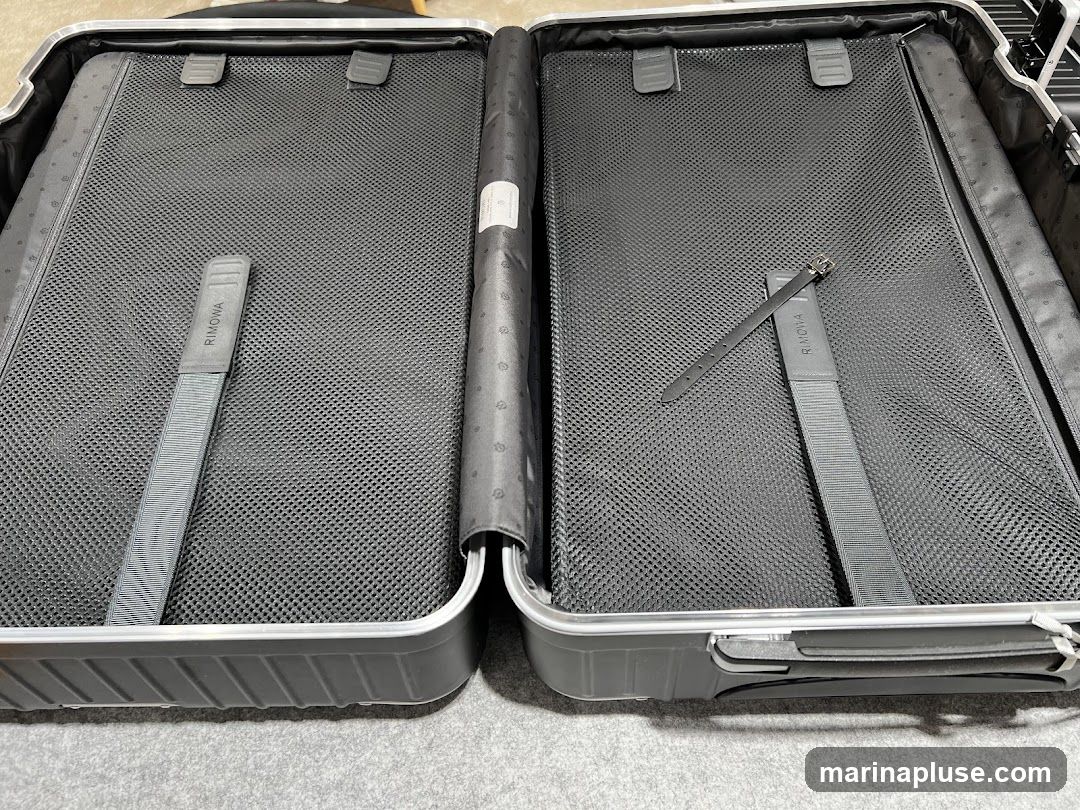 An open Rimowa suitcase, showing the interior compartments. The suitcase is dark gray with silver trim. The interior is lined with black mesh fabric and features two gray straps with the "RIMOWA" logo printed on them. One strap is on the left side, and the other is on the right side, secured with a buckle. The suitcase appears to be empty.