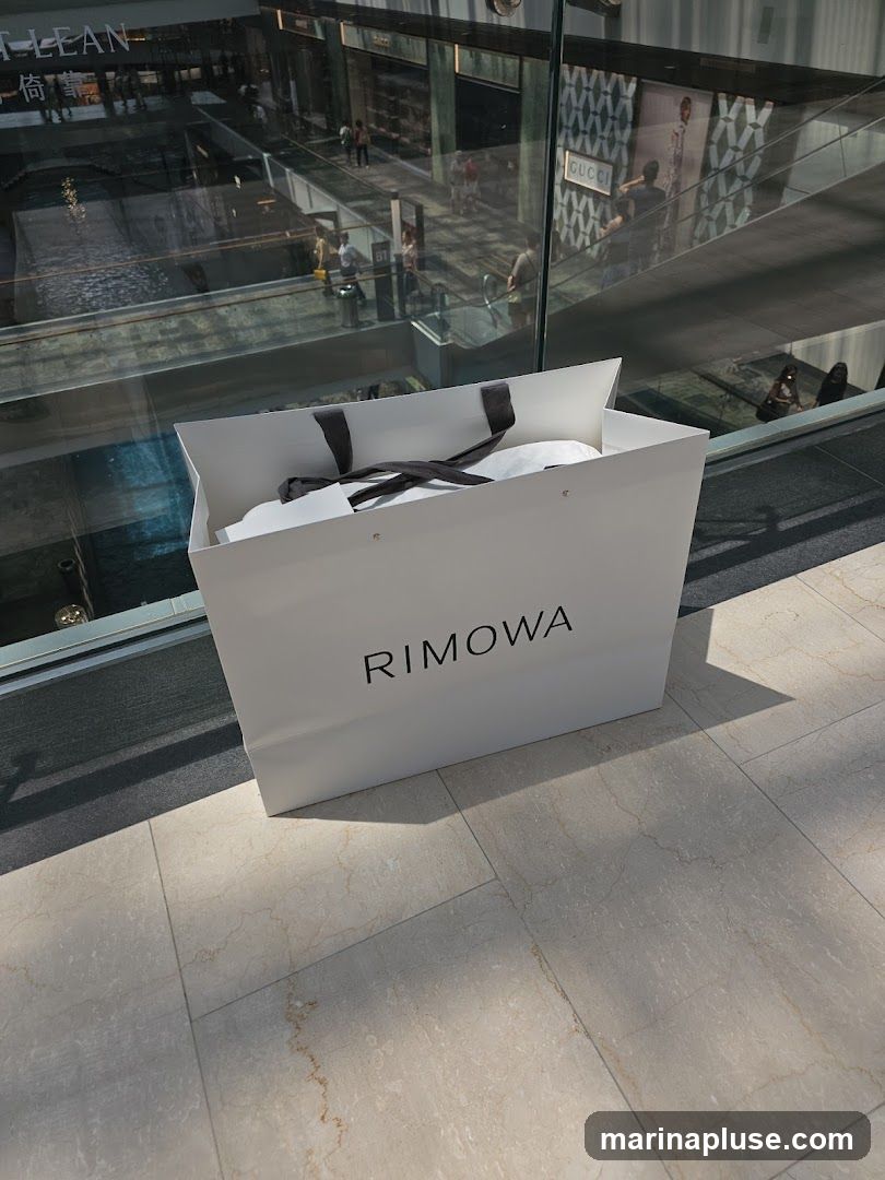 A white shopping bag with the RIMOWA logo printed in black is placed on a tiled floor. The bag is open and appears to contain some white items. In the background, through a glass partition, a multi-level shopping mall interior is visible with escalators, shops, and people. One shop has a prominent GUCCI sign.