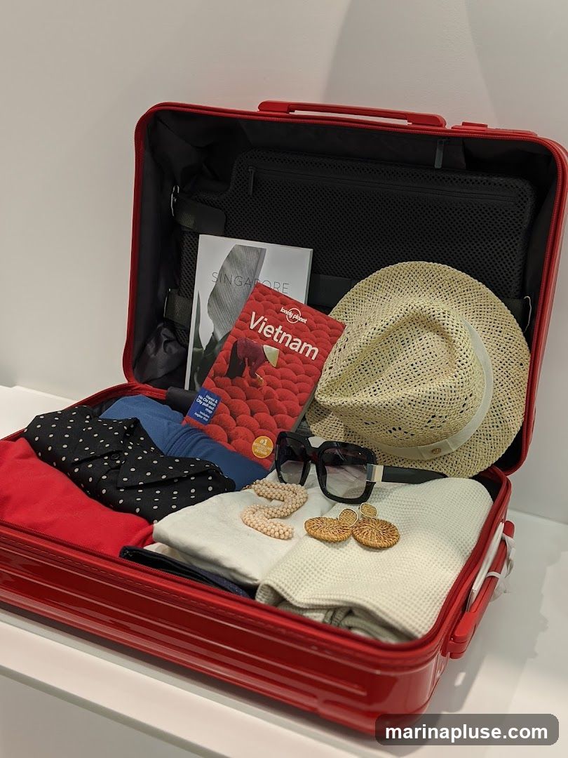 A red suitcase is packed with clothing, a straw hat, sunglasses, jewelry, and travel guides for Singapore and Vietnam.