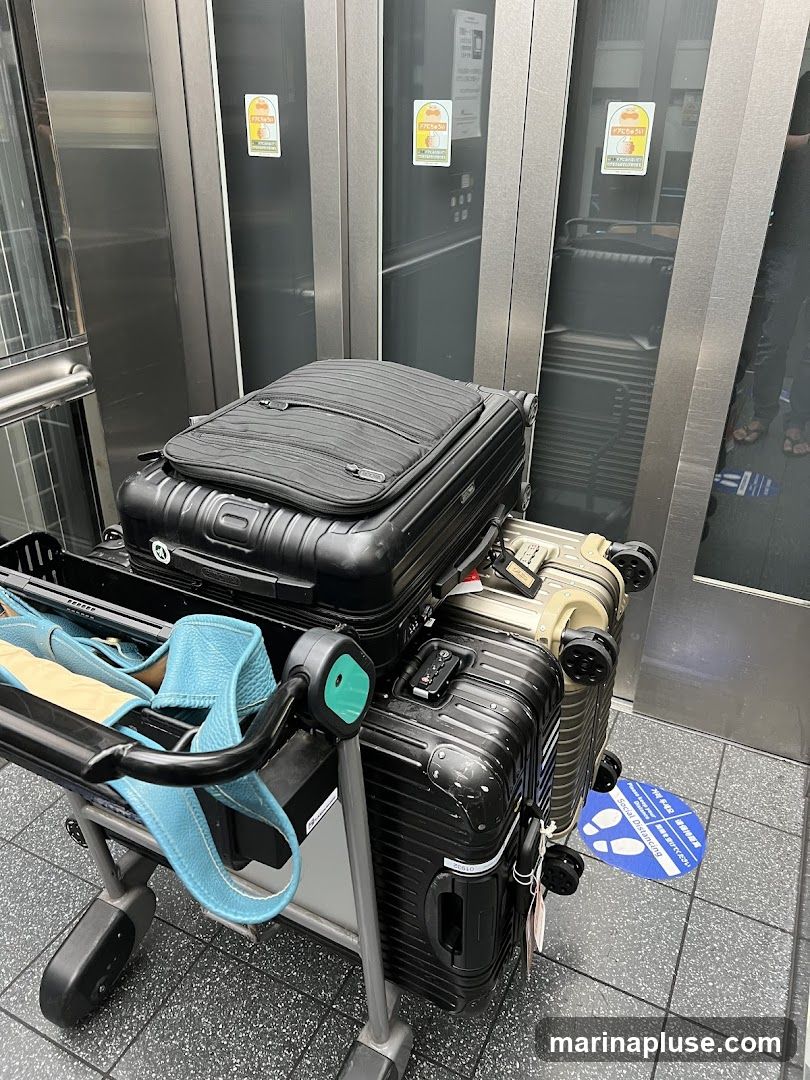 A luggage cart is filled with several suitcases in what appears to be an elevator. The elevator doors are made of reflective metal, and there are yellow stickers with cartoon characters on them. A blue and white floor sticker indicating social distancing is visible on the ground.