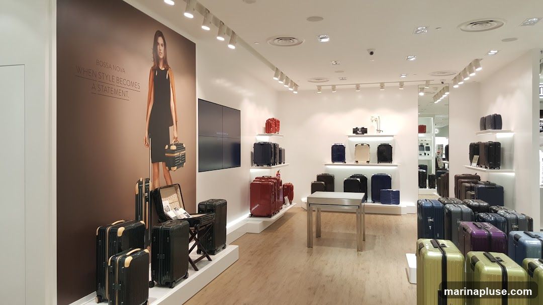 A well-lit interior of a luggage store, showcasing a wide variety of suitcases. A large mural on the left wall features a model with the text "BOSSA NOVA WHEN STYLE BECOMES A STATEMENT." Various suitcases in different colors and sizes are displayed on shelves and on the floor. A metal display table is positioned in the center of the store.