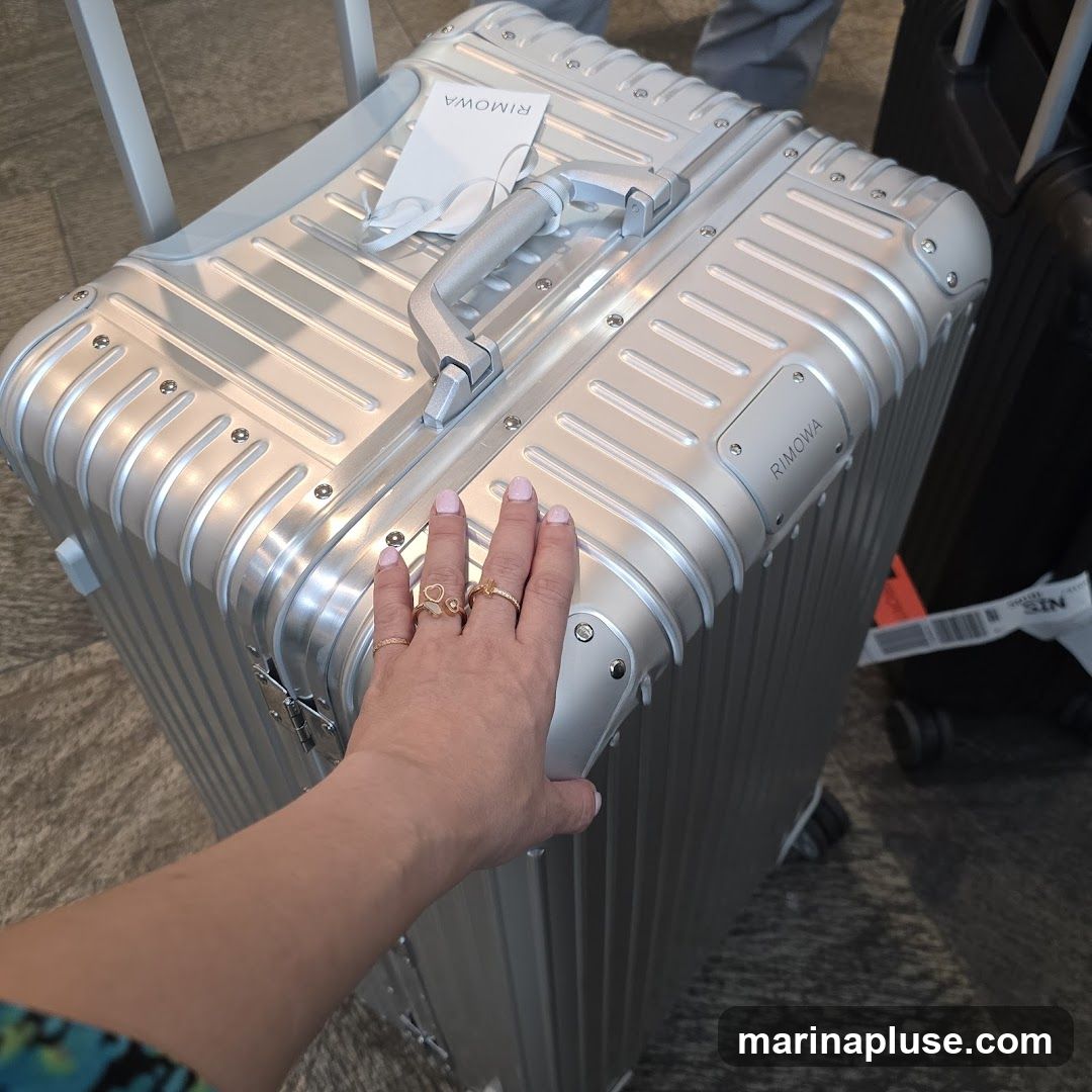 A hand with rings rests on a silver Rimowa suitcase. The suitcase has a textured surface and metal accents. A tag with the Rimowa logo is attached to the handle. Other luggage is visible in the background.