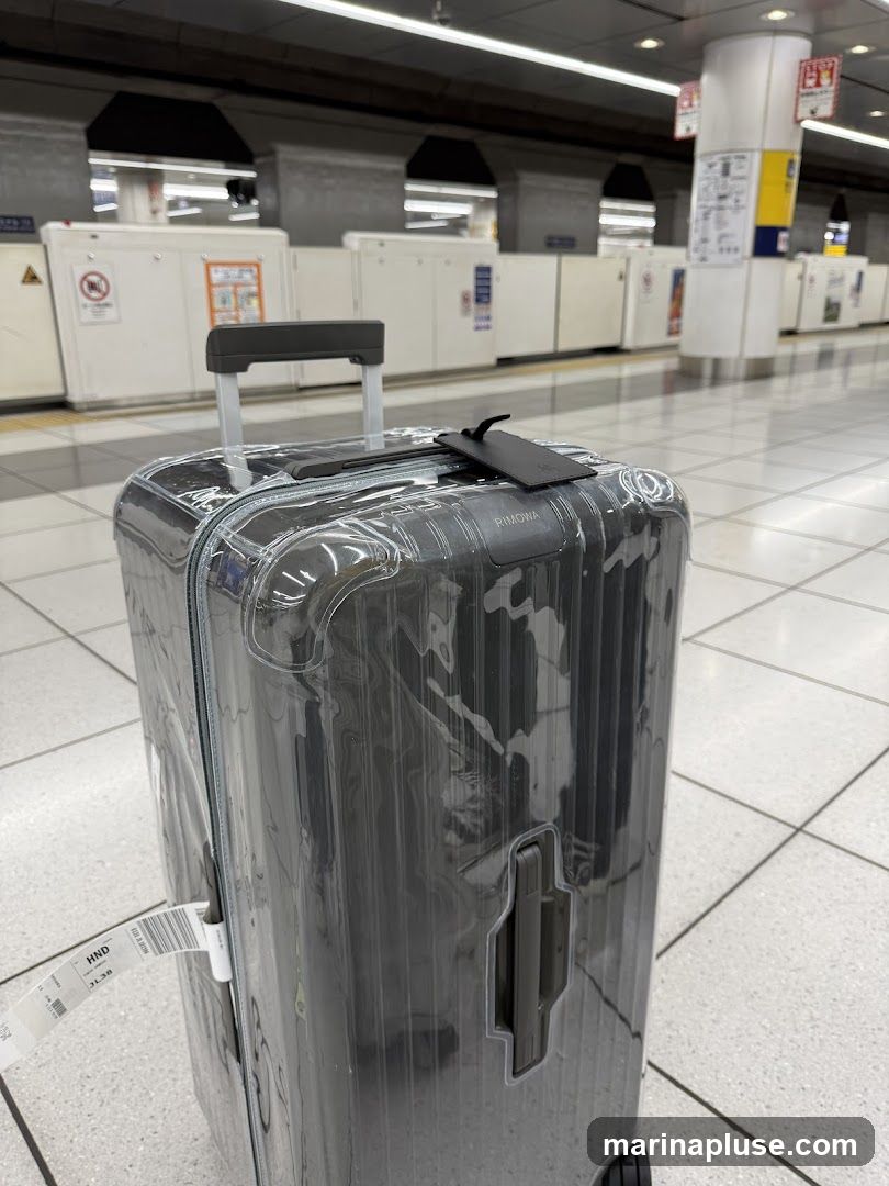 A dark gray Rimowa suitcase covered in clear plastic wrap sits on a tiled floor at a train station. The suitcase has a retractable handle and a black luggage tag. In the background, there are train tracks, platforms, and signage.