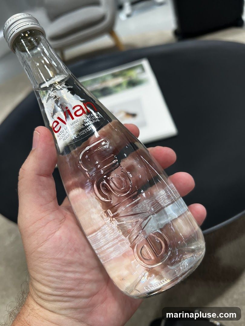 A hand holds a small glass bottle of Evian natural mineral water. The bottle has a silver cap and a red and white label with the Evian logo. The embossed text on the bottle is also visible. In the background, a dark table and a blurred magazine or book can be seen, along with furniture.
