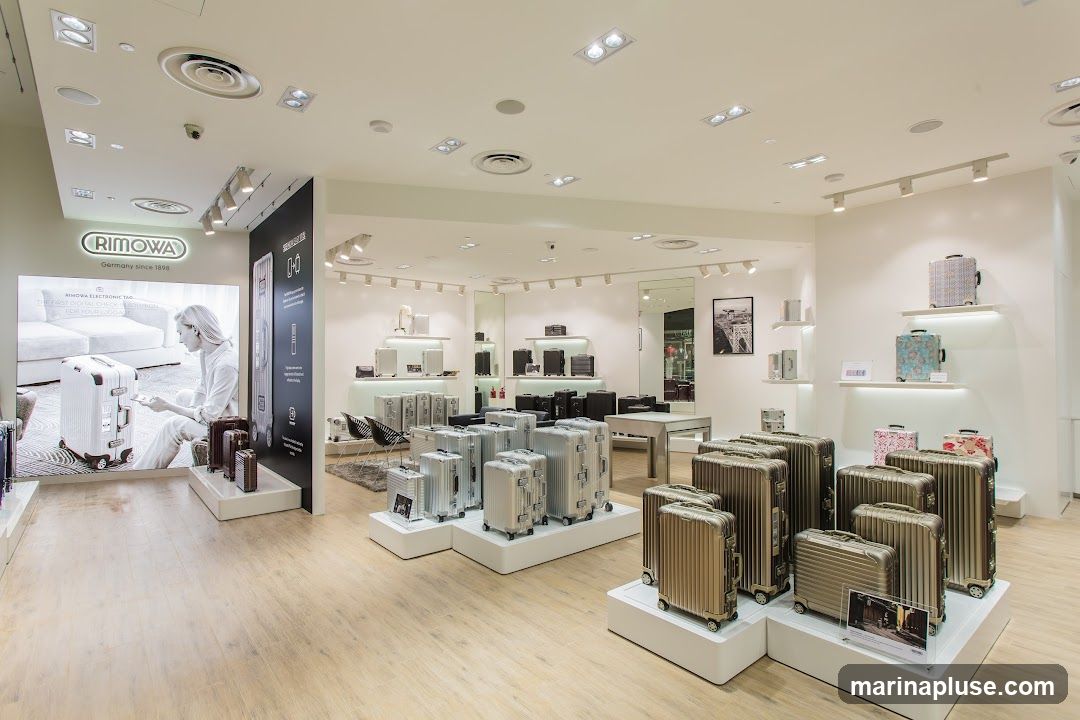 A well-lit interior of a Rimowa store showcasing a variety of their signature aluminum suitcases. The display features numerous silver and gold suitcases of different sizes arranged on white platforms. A large illuminated advertisement on the left wall displays a woman with a Rimowa suitcase and the brand's logo. Shelves on the right wall hold smaller luggage items and decorative pieces. The store has a modern and clean aesthetic with light wooden floors and recessed lighting.