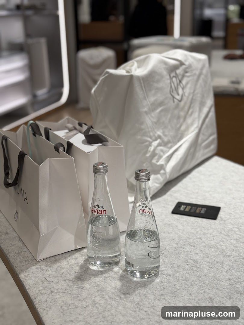 Two glass bottles of Evian water are placed on a grey textured surface next to several white shopping bags. One bag has the word "MOWA" visible. In the background, a large white item covered with a cloth, possibly a piece of furniture or merchandise, is visible. The scene appears to be inside a store or showroom.