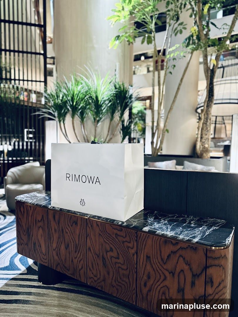 A white Rimowa shopping bag sits on a wooden cabinet with a marble top. The bag has the Rimowa logo printed on it. The background shows an indoor atrium with plants and seating areas.