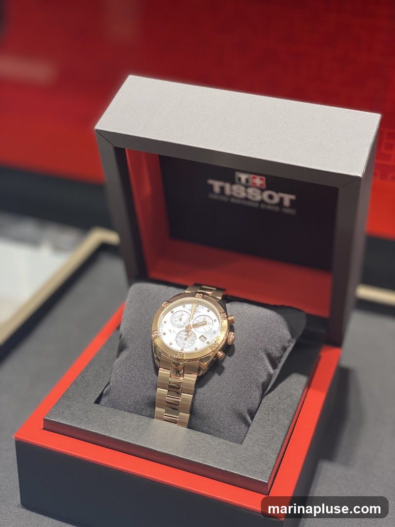 A rose gold Tissot chronograph watch is displayed in its open presentation box. The box has a grey interior with a red trim and a black lid lining featuring the Tissot logo. The watch has a white dial with rose gold hands and markers, and a rose gold metal bracelet.