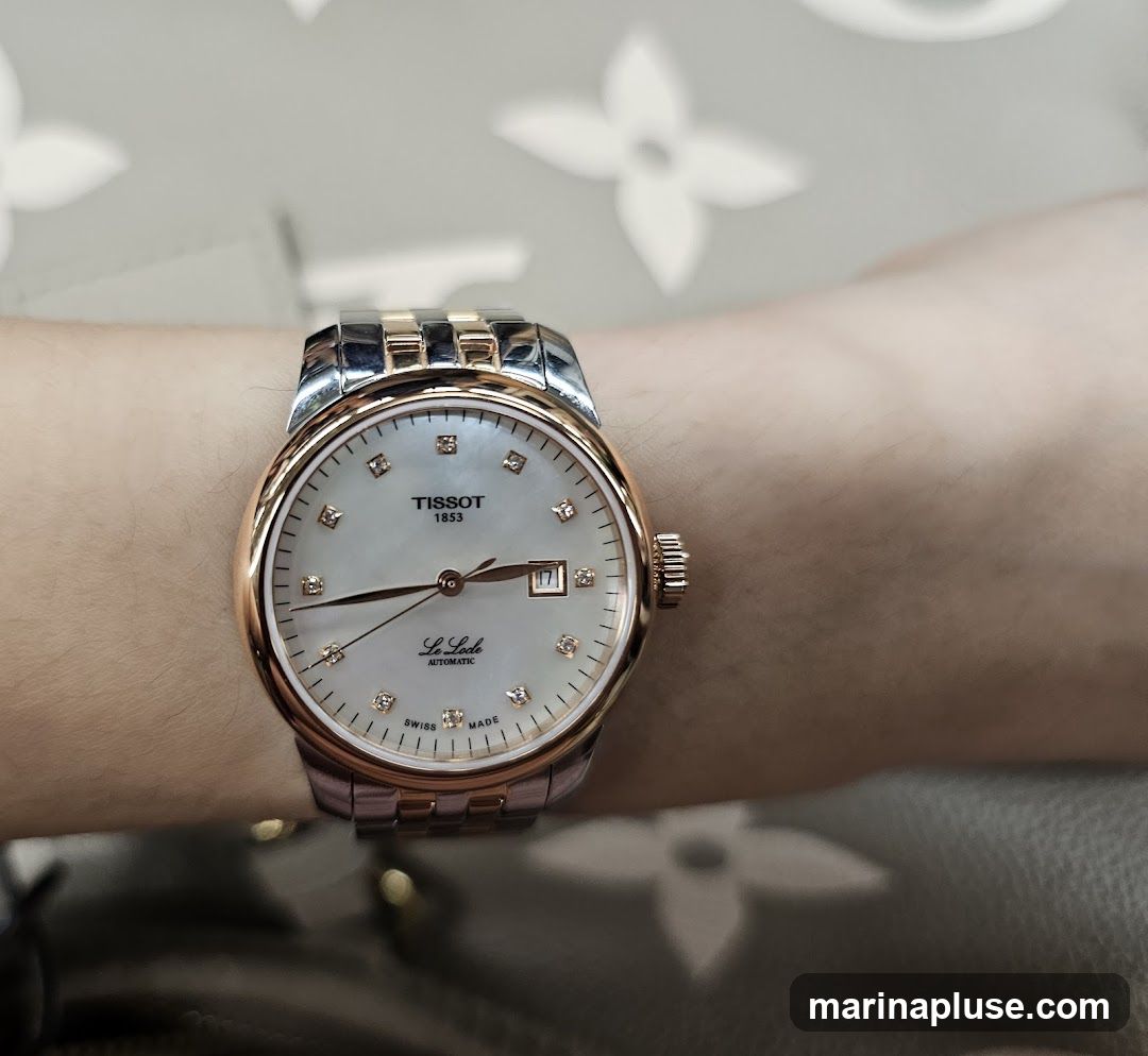 A close-up shot of a Tissot watch on a person's wrist. The watch has a white mother-of-pearl dial with diamond hour markers and rose gold accents. The band is a combination of stainless steel and rose gold. The Tissot logo and "Le Locle Automatic" are visible on the dial.