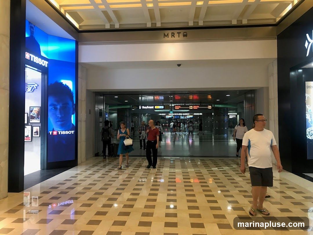 An interior view of a modern shopping mall with a Tissot store on the left featuring a large digital display with a man's face and Tissot branding. In the center, an entrance to an MRT station is visible with signage indicating "Bayfront" and train information. Several people are walking through the mall. The floor is tiled with a checkered pattern, and the ceiling has a grid design.