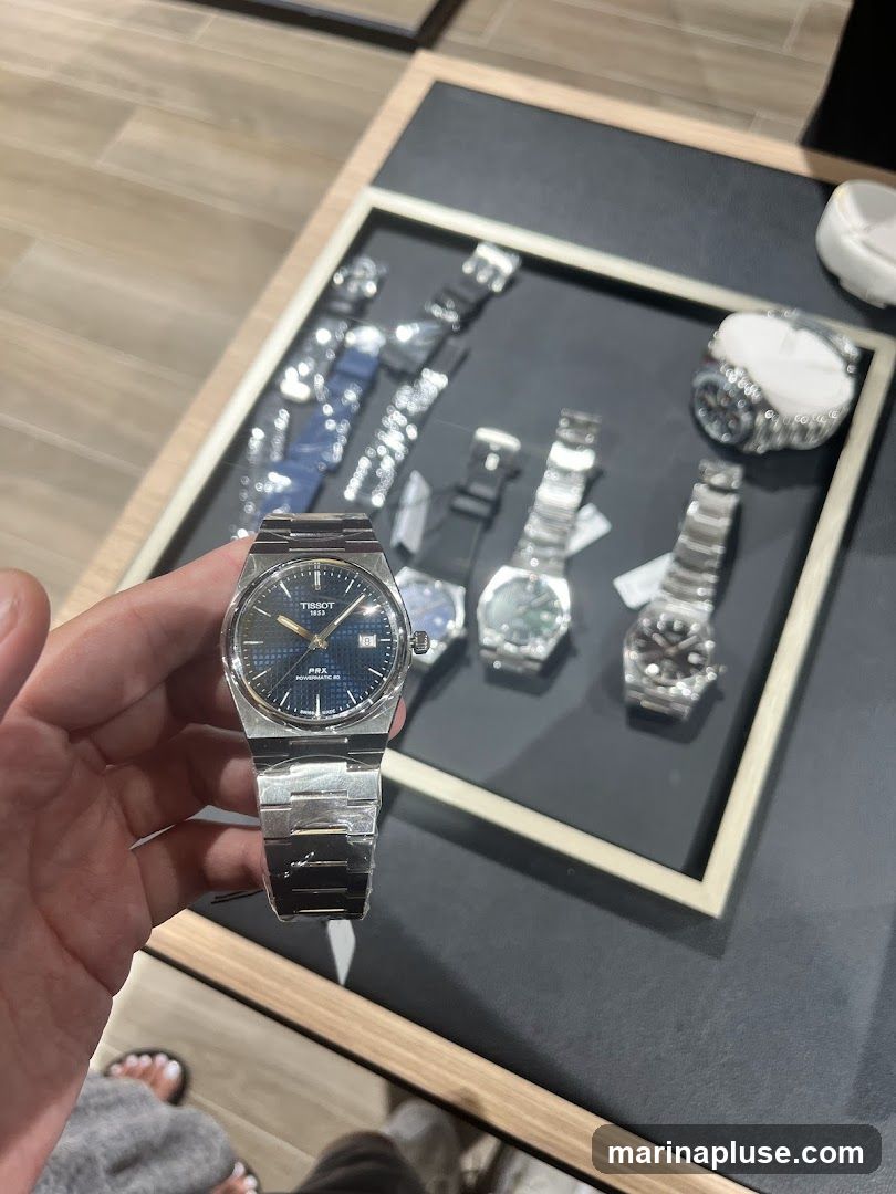 A hand holds a Tissot PRX Powermatic 80 watch with a blue dial. In the background, a display case shows several other watches.