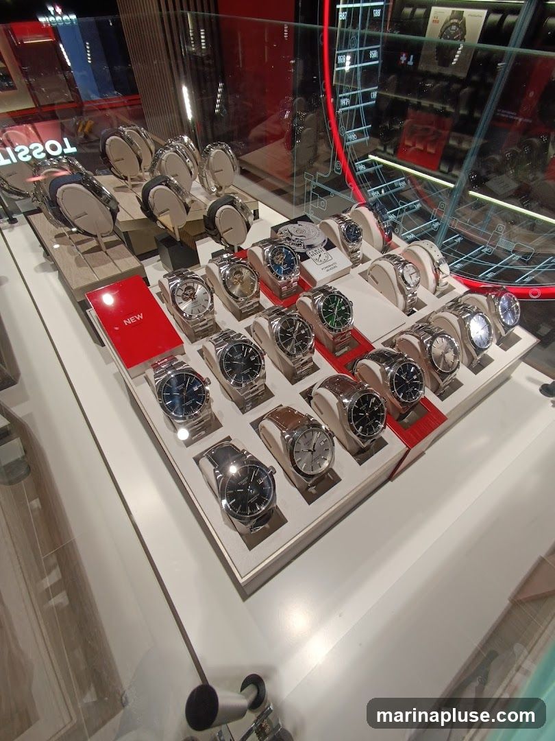 A display case filled with various Tissot watches. The watches are arranged on white stands within a white display tray. Some watches have metal bands, while others have leather bands. A red sign with the word "NEW" is visible on the left side of the display. In the background, more watches and display elements are visible, including a large circular graphic with grid lines and red accents.