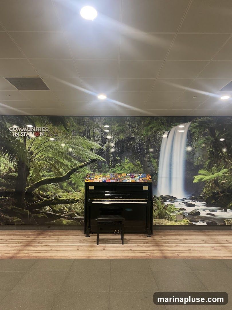 A black upright piano with a bench in front of it is placed in front of a large mural depicting a waterfall and lush greenery. The mural has text that reads "COMMUNITIES IN STATION". The floor in front of the mural is made of wood-patterned tiles, and the floor in front of that is made of grey tiles.