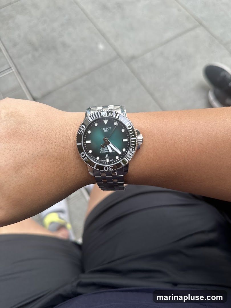 A close-up shot of a person's wrist wearing a Tissot watch with a green dial and a stainless steel band. The watch has a rotating bezel with minute markers. The background shows a tiled surface and blurred legs of a person wearing shorts.