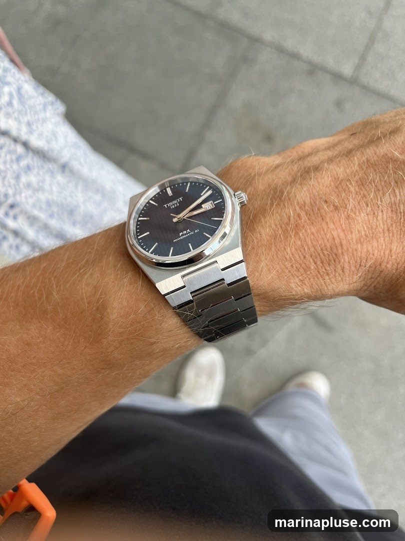 A close-up shot of a Tissot PRX Powermatic 80 watch on a person's wrist. The watch has a dark, textured dial and a stainless steel bracelet. The person is wearing casual clothing, including grey pants and white sneakers. The background appears to be an outdoor setting with a paved surface.